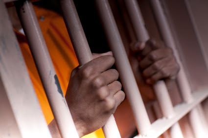A black man with hands outside the bars of a prison cell
